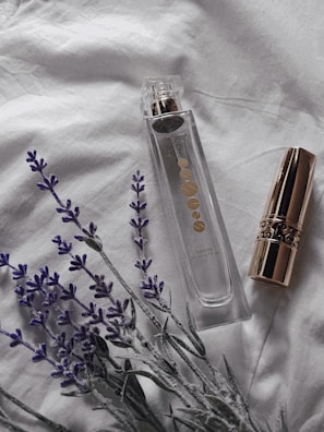 A clear perfume bottle with gold accents is placed on a soft white fabric, accompanied by a gold-capped lipstick and sprigs of lavender. The setting suggests a subtle, elegant atmosphere.