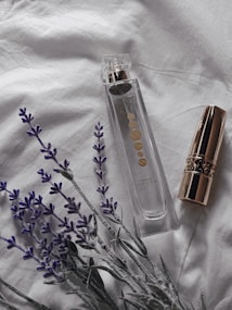 A clear perfume bottle with gold accents is placed on a soft white fabric, accompanied by a gold-capped lipstick and sprigs of lavender. The setting suggests a subtle, elegant atmosphere.