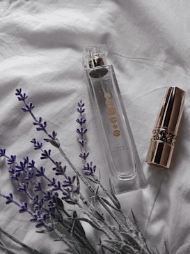 A clear perfume bottle with gold accents is placed on a soft white fabric, accompanied by a gold-capped lipstick and sprigs of lavender. The setting suggests a subtle, elegant atmosphere.