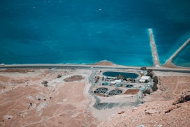 An aerial view of a barren landscape with a large body of blue water and a road running parallel to the shoreline. There is an industrial or utility facility with multiple circular structures and buildings, surrounded by dry, rocky terrain.