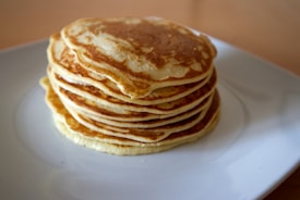 A stack of fluffy, golden-brown pancakes is neatly arranged on a white plate. The pancakes have a light and airy texture, with the top one displaying a slightly darker, crispy edge. The background is softly blurred, emphasizing the pancakes as the central focus.