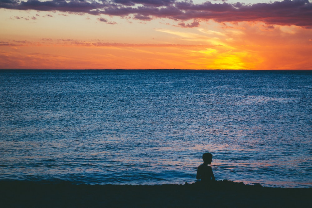 silhouette of man and woman sitting on rock near sea during sunset