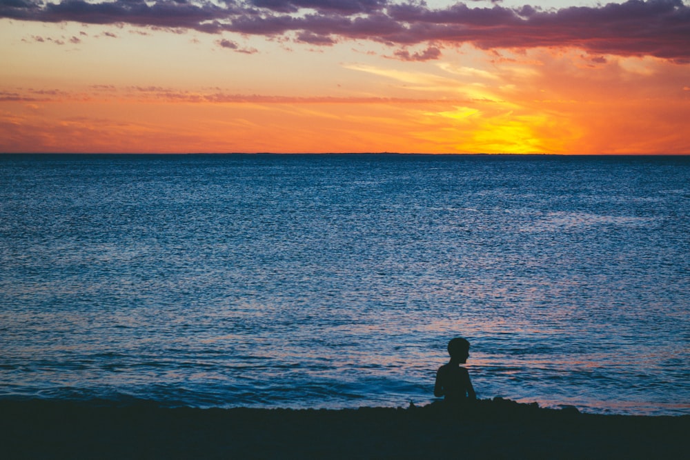 silhueta do homem e da mulher sentados na rocha perto do mar durante o pôr do sol