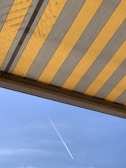 The image features a striped awning with alternating yellow and gray colors, partially open and visible from beneath. In the background, a clear blue sky can be seen with a single white contrail diagonally crossing the frame.