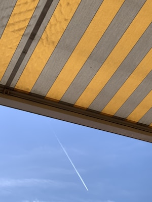 The image features a striped awning with alternating yellow and gray colors, partially open and visible from beneath. In the background, a clear blue sky can be seen with a single white contrail diagonally crossing the frame.