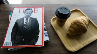 A magazine with a black and white cover featuring a man in a suit is placed on a wooden table. Next to it, there is a wooden tray with a takeaway coffee cup and a croissant.