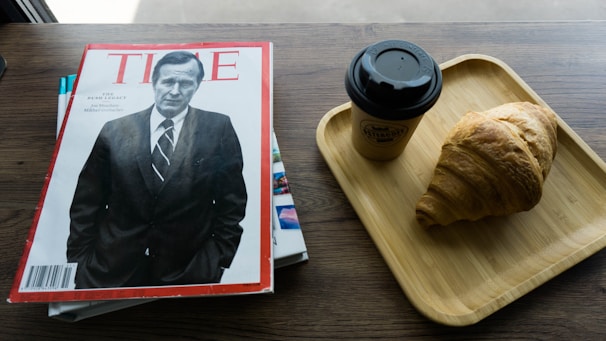 A magazine with a black and white cover featuring a man in a suit is placed on a wooden table. Next to it, there is a wooden tray with a takeaway coffee cup and a croissant.