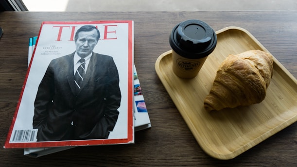 A magazine with a black and white cover featuring a man in a suit is placed on a wooden table. Next to it, there is a wooden tray with a takeaway coffee cup and a croissant.