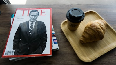 A magazine with a black and white cover featuring a man in a suit is placed on a wooden table. Next to it, there is a wooden tray with a takeaway coffee cup and a croissant.