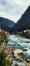 A picturesque mountain river flows through a valley surrounded by lush green hills. On one side, a small village with houses is nestled against the hillside, while on the other side, dense foliage and rocky outcrops frame the river. The sky is overcast, adding a tranquil yet slightly moody ambiance to the scene.