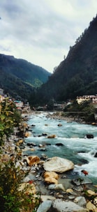 A picturesque mountain river flows through a valley surrounded by lush green hills. On one side, a small village with houses is nestled against the hillside, while on the other side, dense foliage and rocky outcrops frame the river. The sky is overcast, adding a tranquil yet slightly moody ambiance to the scene.