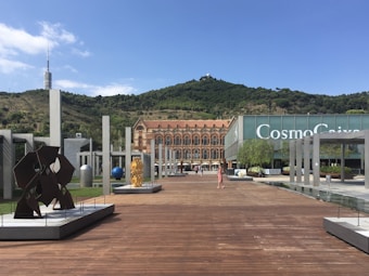 An open plaza with modern art sculptures and a wooden walkway leading to a historic building with ornate architecture. A green hillside serves as the backdrop under a bright blue sky. In the distance, a telecommunications tower is visible.
