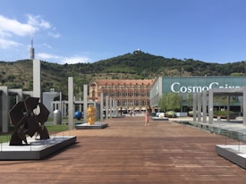 An open plaza with modern art sculptures and a wooden walkway leading to a historic building with ornate architecture. A green hillside serves as the backdrop under a bright blue sky. In the distance, a telecommunications tower is visible.