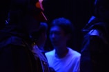 A group of individuals is depicted in a low-light setting, highlighted by deep blue lighting. One person wears a red geometric mask, adding an element of mystery. Their attire suggests a theatrical or artistic context, possibly a performance or rehearsal.