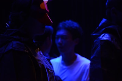 A group of individuals is depicted in a low-light setting, highlighted by deep blue lighting. One person wears a red geometric mask, adding an element of mystery. Their attire suggests a theatrical or artistic context, possibly a performance or rehearsal.