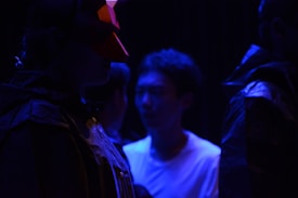 A group of individuals is depicted in a low-light setting, highlighted by deep blue lighting. One person wears a red geometric mask, adding an element of mystery. Their attire suggests a theatrical or artistic context, possibly a performance or rehearsal.