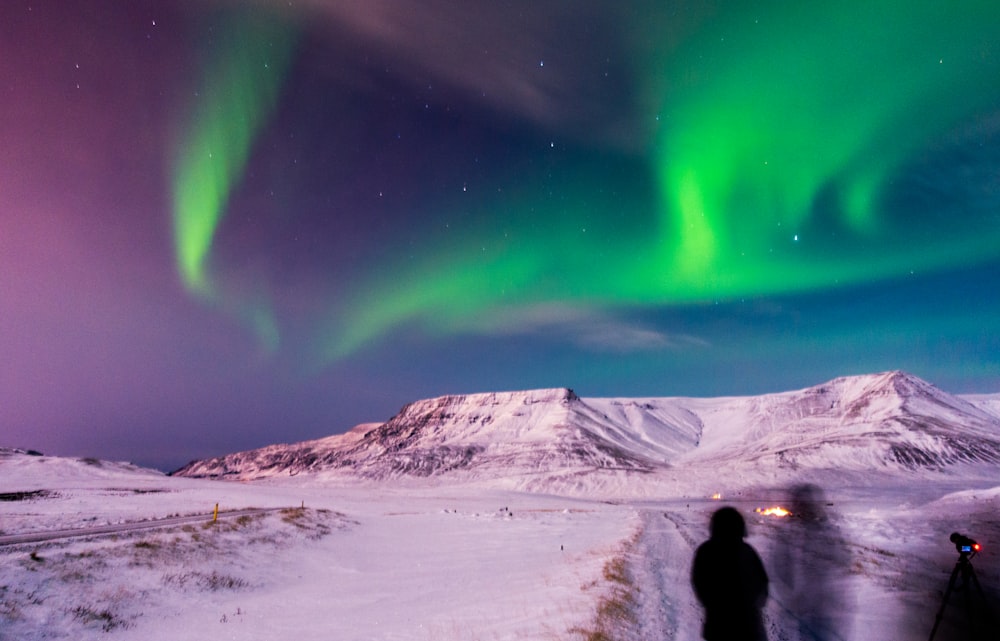 person in black jacket standing on white snow covered field under green aurora lights during night