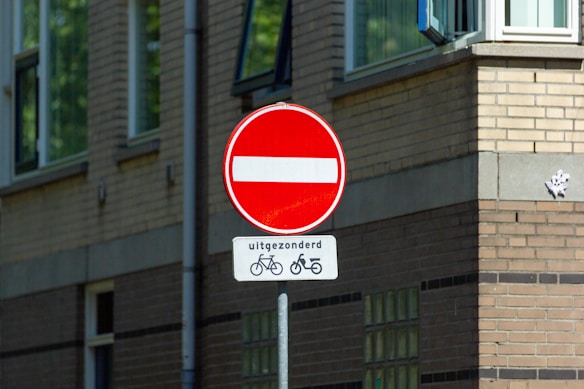 A red no-entry traffic sign with a white horizontal line is displayed prominently. Below it, a smaller sign features Dutch text 'uitgezonderd' alongside symbols indicating exceptions for bicycles and mopeds. The background includes a brick building with several windows, providing an urban setting.