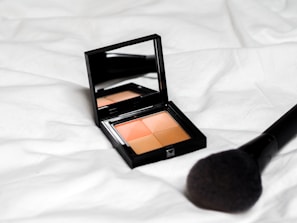 A black makeup compact with a mirror on the inside lid containing four shades of powder sits on a white fabric surface. A large makeup brush with a black handle is placed nearby.
