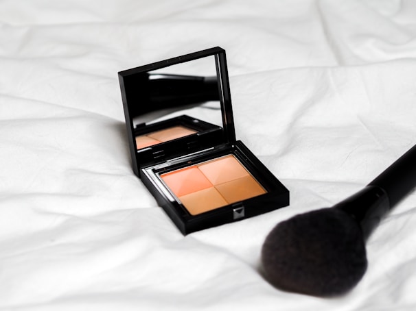 A black makeup compact with a mirror on the inside lid containing four shades of powder sits on a white fabric surface. A large makeup brush with a black handle is placed nearby.