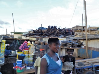 People are walking through a market area carrying items on their heads. A woman in the foreground has a tray of smoked or fried fish balanced on her head, while another person in the background is also carrying goods. The stalls are filled with various products, including colorful containers and packages. The setting appears to be a makeshift outdoor market with corrugated metal and wooden structures.