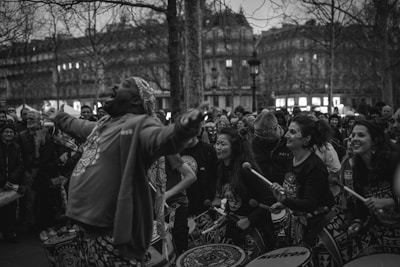 A lively group of drummers and performers is gathered in an open, urban space. The central figure, a man, appears to be singing or shouting enthusiastically with his arms raised. Surrounding him are several other musicians, mostly women, holding drumsticks and smiling. A crowd of onlookers stands behind them, watching the performance, and bare trees and classic architecture form the backdrop as daylight fades.