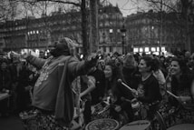 A lively group of drummers and performers is gathered in an open, urban space. The central figure, a man, appears to be singing or shouting enthusiastically with his arms raised. Surrounding him are several other musicians, mostly women, holding drumsticks and smiling. A crowd of onlookers stands behind them, watching the performance, and bare trees and classic architecture form the backdrop as daylight fades.