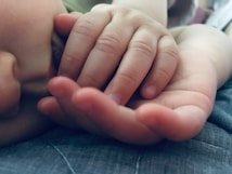 A close-up of a baby's face and hand. The infant's fingers are gently curled, and the skin appears soft and delicate. The image conveys a sense of tenderness and warmth.