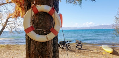A life preserver hangs on a tree near the sandy shoreline of a lake. In the background, the blue water stretches to the horizon under a clear sky. On the beach, two wooden chairs and a table face the water, and a yellow kayak rests on the sand.