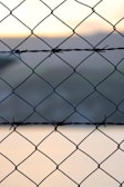A close-up view of a chain-link fence made of metal wire. The fence features diamond-shaped patterns with knots and some barbed wire intertwined. The background is blurred, showcasing soft hues of sunset or sunrise lighting.