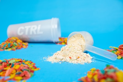 A plastic shaker bottle labeled 'CTRL' is lying on its side. In front of it, there is a white plastic scoop filled with a powder that resembles a nutritional or protein supplement. Around the scoop and bottle, colorful cereal pieces are scattered on a bright blue background.