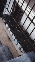 A wet concrete surface with puddles is bordered by metal railings. The scene captures the reflection of the railings in the water on the ground, creating a pattern. The area appears to be a walkway or a stair landing with a slightly gloomy atmosphere.