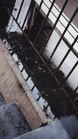A wet concrete surface with puddles is bordered by metal railings. The scene captures the reflection of the railings in the water on the ground, creating a pattern. The area appears to be a walkway or a stair landing with a slightly gloomy atmosphere.