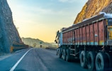 A large truck is parked on a winding road bordered by steep rocky cliffs. The scene is bathed in the warm glow of the setting or rising sun, casting a golden hue on the landscape. The road is mostly empty, stretching into the distance with safety barriers on one side.