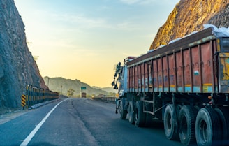 A large truck is parked on a winding road bordered by steep rocky cliffs. The scene is bathed in the warm glow of the setting or rising sun, casting a golden hue on the landscape. The road is mostly empty, stretching into the distance with safety barriers on one side.