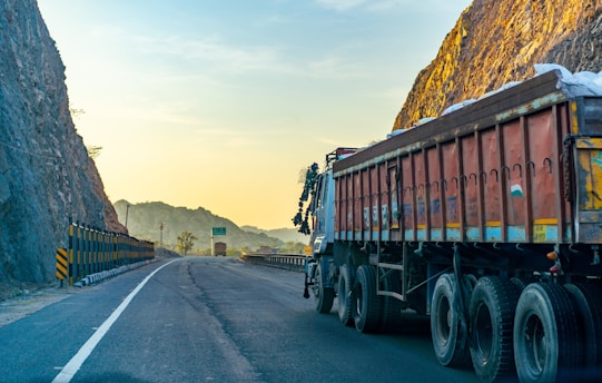 A large truck is parked on a winding road bordered by steep rocky cliffs. The scene is bathed in the warm glow of the setting or rising sun, casting a golden hue on the landscape. The road is mostly empty, stretching into the distance with safety barriers on one side.