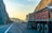 A large truck is parked on a winding road bordered by steep rocky cliffs. The scene is bathed in the warm glow of the setting or rising sun, casting a golden hue on the landscape. The road is mostly empty, stretching into the distance with safety barriers on one side.