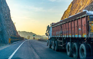 A large truck is parked on a winding road bordered by steep rocky cliffs. The scene is bathed in the warm glow of the setting or rising sun, casting a golden hue on the landscape. The road is mostly empty, stretching into the distance with safety barriers on one side.
