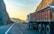 A large truck is parked on a winding road bordered by steep rocky cliffs. The scene is bathed in the warm glow of the setting or rising sun, casting a golden hue on the landscape. The road is mostly empty, stretching into the distance with safety barriers on one side.