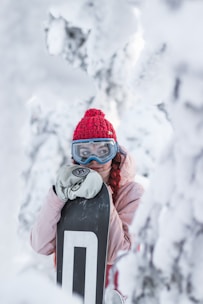 A person dressed in winter attire stands in a snowy setting, holding a snowboard. They wear a red knit hat, blue ski goggles, and a light pink jacket. The surroundings are covered in snow, creating a serene winter landscape.