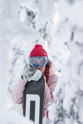 A person dressed in winter attire stands in a snowy setting, holding a snowboard. They wear a red knit hat, blue ski goggles, and a light pink jacket. The surroundings are covered in snow, creating a serene winter landscape.