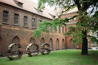 A historical building with a brick facade featuring arched windows and a sloped roof. In front, a large metal sculpture spells out the letters 'SOS' on a lawn, surrounded by dense green foliage from a nearby tree.