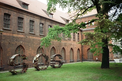 A historical building with a brick facade featuring arched windows and a sloped roof. In front, a large metal sculpture spells out the letters 'SOS' on a lawn, surrounded by dense green foliage from a nearby tree.