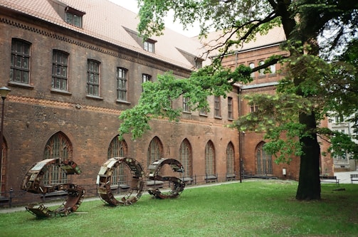 A historical building with a brick facade featuring arched windows and a sloped roof. In front, a large metal sculpture spells out the letters 'SOS' on a lawn, surrounded by dense green foliage from a nearby tree.