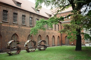 A historical building with a brick facade featuring arched windows and a sloped roof. In front, a large metal sculpture spells out the letters 'SOS' on a lawn, surrounded by dense green foliage from a nearby tree.
