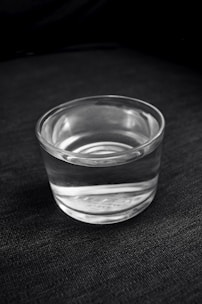 A clear glass filled with water is placed on a dark, textured surface. The transparency of the glass and the reflection of light create subtle highlights and shadows.