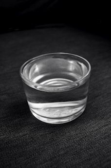 A clear glass filled with water is placed on a dark, textured surface. The transparency of the glass and the reflection of light create subtle highlights and shadows.