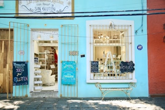 A bright turquoise storefront features a welcoming entrance with a sign overhead advertising a gluten-free kitchen. The open door reveals a cozy interior with shelves and decorative furnishings. A large window displays various items, and two blackboards hang on either side of the entrance, offering menus and promotions. A decorative bench sits in front of the building.