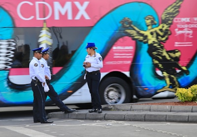 Three uniformed officers stand on the street in front of a colorful bus featuring bold graphics and the text 'CDMX'. The bus design includes a stylized golden statue and turquoise swirl against a pink background. The officers, two standing together and one separate, appear to be engaged in conversation, with the lone officer holding a notepad.