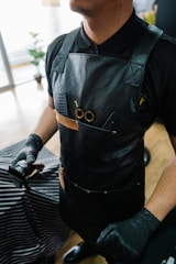 A barber wearing a black leather apron and black gloves stands with scissors and a comb in the apron pocket. The setting includes a striped barber cape over a customer and a wooden floor.